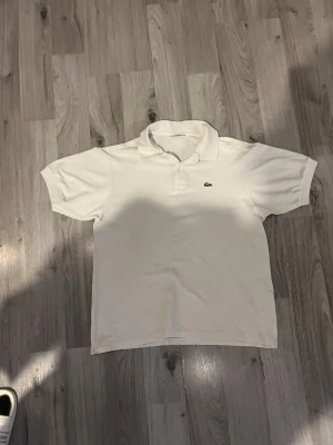 Vit piké från Lacoste - Klassisk vit pikétröja från Lacoste med korta ärmar och den ikoniska krokodilloggan på bröstet. Tröjan har krage och knappar vid halsen. Perfekt för en clean och stilren look och den har en liten fläck i magen men det syns inte så mycket.