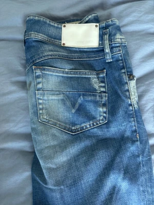 Blå slitna jeans från Diesel - Säljer ett par blå jeans från Diesel med slitningar och tvättade detaljer. Klassisk femficksmodell med låg midja och raka ben. Jeansen har en vit patch bak i midjan och diskreta logodetaljer. Perfekta för en avslappnad och trendig look.