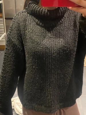 Svart grovstickad polotröja - Mysig svart grovstickad polotröja med hög krage och avslappnad passform. Tröjan har långa ärmar och ribbstickade muddar vid halsen. Perfekt för kalla dagar och enkel att matcha med olika outfits. Tröjan har även små glittriga flätade detaljer på sidan av armarna och upp till nacken.