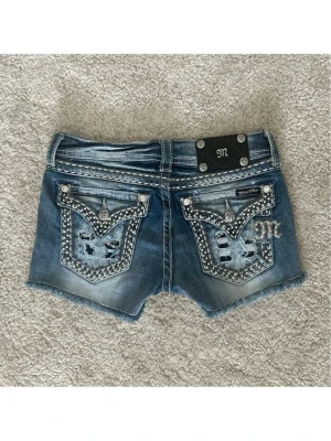 Miss Me blå jeansshorts med strass - Snygga blå jeansshorts från Miss Me med slitna detaljer, råa kanter och blingade bakfickor med strass och broderier. Shortsen har låg midja, dekorativa sömmar och coola nitar. Perfekta för dig som vill sticka ut med en edgy och glam stil. Midjemått: 72cm 💞