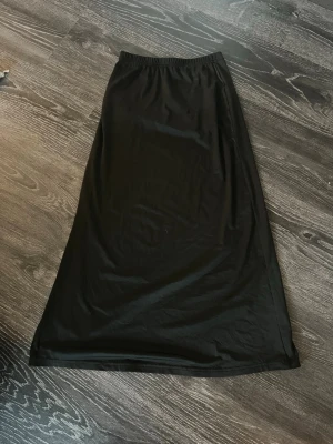 Svart långkjol med slits - En svart långkjol med elastisk midja och slits på sidan. Kjolen har en enkel och stilren design som passar till många olika looks. Tillverkad i ett mjukt och lätt material som ger en skön känsla.