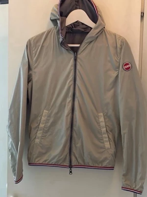 Beige vindjacka från Colmar - Stilren beige vindjacka från Colmar med huva och dragkedja framtill. Jackan har två snedställda fickor och Colmar-logga på ärmen. Detaljer i rött och vitt vid muddar och nederkant. Tillverkad i lätt, vindtätt material som passar perfekt till vår och höst. Ny pris 2700kr