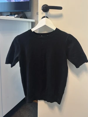 Svart kortärmad tröja - En stilren svart tröja med korta ärmar och rund hals. Tröjan har en ribbad kant vid halsen och är tillverkad i ett mjukt material som passar perfekt till många olika outfits. 