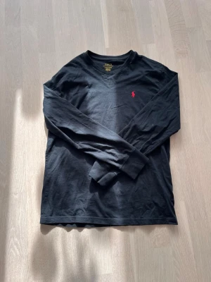 Svart långärmad tröja från Polo Ralph Lauren - Klassisk svart långärmad tröja från Polo Ralph Lauren med röd broderad logga på bröstet. Tröjan har v-ringning och är gjord i mjuk bomull. Den är använd fåtal gånger. Skick-8/10.  Den är L 14-16 som är som ungefär en XS. Passar runt 165-175. Tveka inte att fråga om referensbilder! 