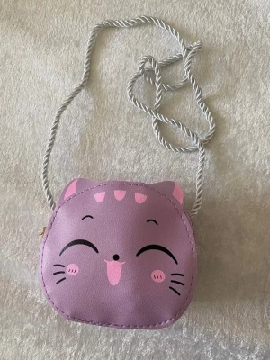 Söt kattväska  - Två små handväskor i form av glada kattansikten, en i lila och en i ljusrosa. Väskorna har vita snoddar som axelband och är gjorda i konstläder med tryckt kattmotiv och öron. Perfekt för dig som gillar gulliga accessoarer och vill sticka ut.  69kr/st 