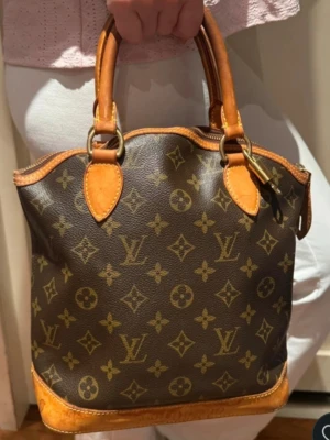 Louis Vuitton Monogram handväska, lock it leather handbag. - Säljer en klassisk Louis Vuitton handväska i brunt monogrammönstrat canvas med detaljer i ljust brunt läder. Väskan har två stadiga handtag och guldfärgade metalldetaljer. Ikoniskt LV-mönster över hela väskan och snygg vintagekänsla. Hänglås med nyckel. Fläckar finns på lädret undersida. Kod finns samt verifierad. 