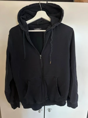 Svart zip hoodie med fickor - Klassisk svart hoodie med dragkedja från Dressman. Tröjan har huva med dragkedja, två framfickor och ribbade muddar vid ärmar och nederkant. Perfekt för en avslappnad och enkel stil. Tillverkad i mjuk bomullsblandning. Dock är ena änden till snöret borta men inget man lägger märke till! Storlek S