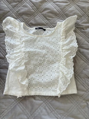 Vit blus med volang från Zara - Supersöt vit blus från Zara med broderade detaljer och stora volanger framtill. Blusen har rund halsringning och är ärmlös, perfekt för en fräsch och somrig look. Materialet känns lätt och luftigt, och den broderade spetsen ger en romantisk vibe.