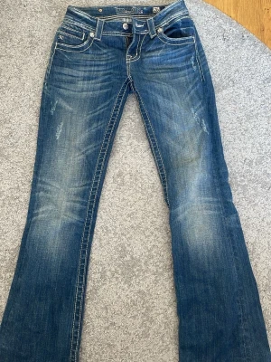 Blå bootcut jeans från Miss Me - Säljer ett par blå bootcut jeans från Miss Me med slitningar och markerade vita sömmar. Jeansen har låg midja, fem fickor och råa, fransiga benslut. Materialet är klassisk denim i bomull med snygg tvätt och detaljerade sömmar. Sydde in midjan så storleken är ej storlek 26 som det står i byxorna men mått finns Bladins bilderna. Köpta här på Plick och förra ägaren klippte av en bit vid ändrarna. Innerbenslängden är ca 73 cm