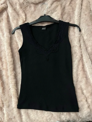 Svart ribbad linnetopp med spets - Svart ribbad linnetopp från Gina Tricot med v-ringad spetsdetalj vid halsen. Ärmlös och figurnära passform, perfekt för att styla med jeans eller kjol. Materialet är mjukt och stretchigt för en bekväm känsla.