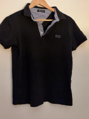 Svart pikétröja från Hugo Boss - Klassisk svart pikétröja från Hugo Boss med korta ärmar och vitrandig insida på kragen. Tröjan har en liten logga broderad på bröstet och stängs med två knappar vid halsen. Perfekt till jeans eller chinos.