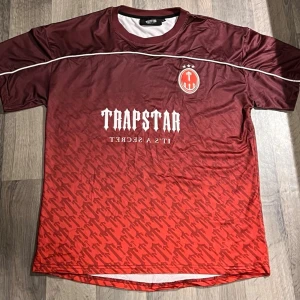 Röd Trapstar football Jersey - Snygg fotbollströja från Trapstar i röd och vinröd färg med vit logga och text. Tröjan har ett mönster nertill, tryckt nummer 22 på ryggen och rund hals. Perfekt för dig som gillar streetwear och sportig stil.