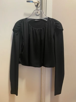 Svart långärmad topp från Gina Tricot - Svart långärmad topp från Gina Tricot med rynkade axlar och rund halsringning. Modellen är croppad och har en stilren look som passar till många outfits. Tillverkad i ett mjukt och följsamt material.