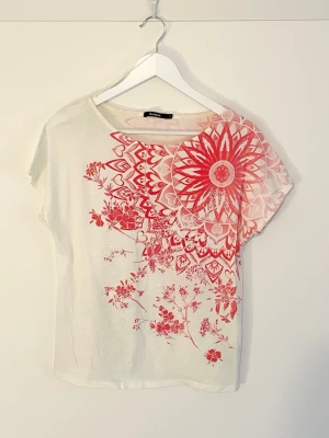 Mönstrad topp från Desigual - Snygg vit t-shirt från Desigual med korta ärmar och stort rött blommönster samt mandalainspirerat tryck över axeln. T-shirten har en avslappnad passform och är gjord i mjuk bomull, perfekt för en trendig vardagslook. Tröjan har kvar etikett med tvätt instruktioner och har bara använts enstaka gånger. Originalpris 699 kr.
