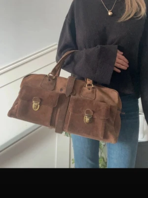 Brun mockaväska med fickor - Säljer en snygg handväska i brun mocka med två stora framfickor och guldfärgade spännen. Väskan har ett klassiskt rektangulärt format och ett handtag upptill. Perfekt för dig som gillar vintagevibbar och praktiska detaljer.