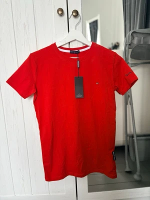 Röd t-shirt från Tommy Hilfiger - Snygg röd t-shirt från Tommy Hilfiger med klassisk passform och rund hals. Liten logga på bröstet och broderad text på ärmen. Tillverkad i mjuk bomull som känns skön mot huden. Perfekt till jeans eller shorts för en clean look.