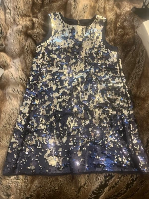 Glittrig blå kortklänning med paljetter - Snygg ärmlös kortklänning i mörkblått med massor av silverfärgade paljetter som ger ett riktigt glittrigt intryck. Klänningen har en rak och enkel form, perfekt för dig som vill sticka ut. Materialet är polyester och klänningen har en liten knapp i nacken.