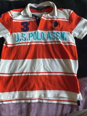 Randig pikétröja från U.S. Polo Assn. - Snygg randig pikétröja från U.S. Polo Assn. i orange och vitt med blå broderad logga och text på bröstet. Tröjan har klassisk krage, korta ärmar och knappar vid halsen. Perfekt för en sportig och avslappnad stil.