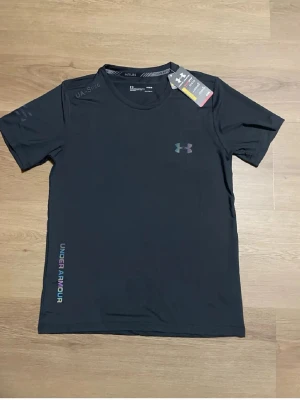 Svart tränings-tshirt Under Armour - Snygg svart tränings-tshirt från Under Armour med rund hals och korta ärmar. Diskret logga på bröstet och texten UNDER ARMOUR längs sidan. Tillverkad i lätt och snabbtorkande funktionsmaterial, perfekt för löpning eller gym.