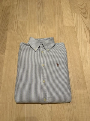 Randig skjorta från Ralph Lauren XS - Klassisk blå- och vitrandig skjorta från Ralph Lauren i modellen Custom Fit. Skjortan har knappar framtill, button-down krage och den ikoniska broderade loggan på bröstet. Tillverkad i mjuk bomull och passar perfekt till en clean och stilren look.