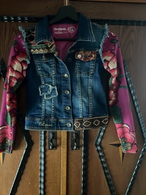 Desigual jeansjacka med mönstrade ärmar - Unik jeansjacka från Desigual med broderade detaljer, patchwork och dekorativa knappar. Ärmarna är i rosa och lila med stora blommönster och kontrasterar snyggt mot den blå denimkroppen. Fransen vid axlarna och mixade material ger jackan en färgstark och kreativ look.