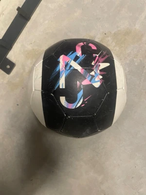 Svartvit fotboll med färgglad logga - Säljer en svartvit fotboll med en stor färgglad logga i blått, rosa och vitt på ena sidan. Bollen har klassiskt mönster med svarta och vita fält och är perfekt för spontanfotboll eller träning.