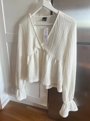 Vit omlottblus från Gina Tricot - Helt ny vit blus från Gina Tricot i ett strukturerat, krinklat tyg. Blusen har långa, puffiga ärmar med resår och volang vid ärmslut samt en avslappnad passform och djup V-ringning. 
