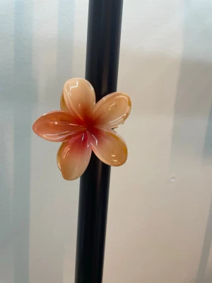 Blommig hårklämma i persikofärg - Söt hårklämma formad som en blomma i persikofärgade och röda toner. Klämman är gjord i plast med glansig finish och har fem kronblad. Perfekt för att ge en färgklick till din frisyr och hålla håret på plats.