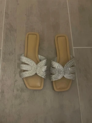 Glittriga slip-in sandaler med strass - Säljer ett par snygga slip-in sandaler med fyrkantig tå och breda band täckta av silverfärgad strass. Sulan är beige och har en mjuk look. Perfekta för dig som vill ha något extra till outfiten. Märke ej synligt.