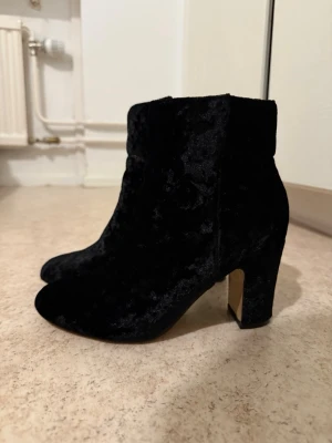 Svarta sammetsstövletter med klack - Säljer ett par svarta stövletter i mjuk sammet med chunky klack och rundad tå. Skorna har en stilren siluett och passar perfekt till höstens och vinterns outfits. Inga synliga detaljer eller loggor, vilket ger en clean och trendig look.