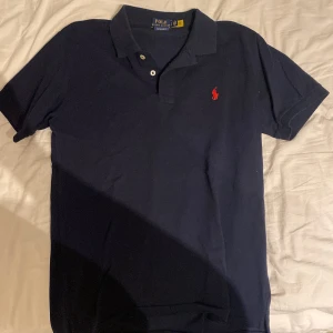Marinblå pikétröja från Polo Ralph Lauren - Klassisk marinblå pikétröja från Polo Ralph Lauren med röd broderad logga på bröstet. Tröjan har korta ärmar, krage och knappar vid halsen. Modellen är custom slim fit och passar perfekt till en avslappnad men stilren look.