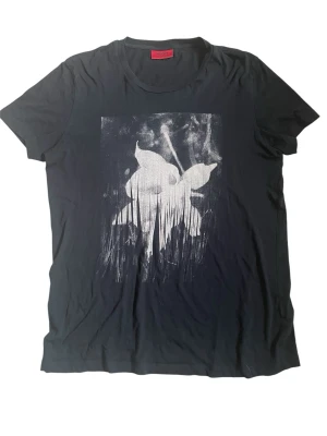 Svart t-shirt med print från Hugo Boss - Cool svart t-shirt från Hugo Boss med unikt beige print på framsidan. T-shirten har korta ärmar och är tillverkad i mjuk bomull. Perfekt för dig som vill ha en stilren men edgy look.