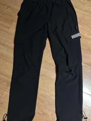 Svarta cargobyxor från Hoodrich - Säljer ett par svarta cargobyxor från Hoodrich med tryckt logga på benet och textdetaljer baktill. Byxorna har flera fickor, dragsko i benslut och elastisk midja. Perfekta för en streetwear-look och riktigt sköna att bära till vardags. Slitet textdetaljer baktill.