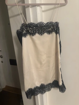 Krämvit spetskantad linnetopp - Snygg krämvit linnetopp med tunna axelband och breda svarta spetsdetaljer längs både över- och nederkant. Toppen har en rak passform och ger en elegant och trendig look. Perfekt att styla med jeans eller kjol för en chic outfit.