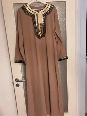 Beige långklänning med paljettdetaljer - Säljer en beige långklänning från My Style med långa ärmar och vacker paljettdekor i guld och svart runt halsringningen och ärmsluten. Klänningen har en rak passform och ger en elegant look med sina skimrande detaljer.