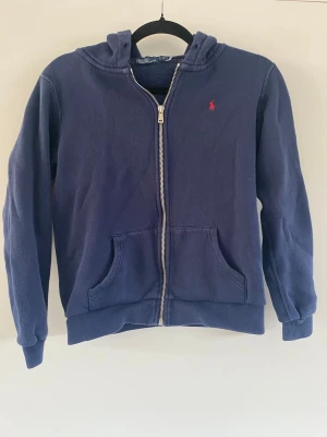 Marinblå hoodie från Polo Ralph Lauren - Klassisk marinblå hoodie med dragkedja från Polo Ralph Lauren. Hoodien har huva, två fickor framtill och ribbade muddar. Liten röd logga broderad på bröstet. Saknar tyvärr snören, men går lätt att sätta i om man så önskar!