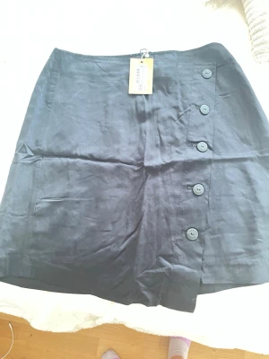 Svart kortkjol med knappar - Snygg svart kortkjol i satinliknande material med dekorativa knappar längs sidan. Kjolen har en clean look och passar perfekt till många olika stilar. Enkel att matcha och supertrendig.