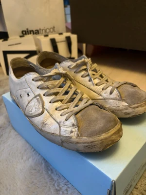 Philippe Model, Vita låga sneakers med grå detaljer - Säljer ett par vita sneakers med grå detaljer och slitna kanter. Skorna har låga skaft, snörning och en klassisk siluett. Materialet är en mix av läder och textil. Perfekta för en avslappnad streetstyle-look.