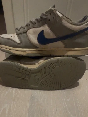 Nike Dunk Low grå/blå sneakers - Nike Dunk Low sneakers i grått och vitt läder med blå Swoosh och detaljer. Klassisk siluett med perforerad tåbox och grå snörning. Yttersulan är beige och har retro-vibe. Perfekta för dig som gillar streetwear och tidlös design.