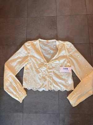 Gul broderad blus från Nelly - Söt gul blus från Nelly med broderade blommor och hålmönster. Blusen har långa ärmar med resår vid ärmslut, v-ringning och knytband framtill. Tillverkad i ett lätt och luftigt material som passar perfekt till sommaren.