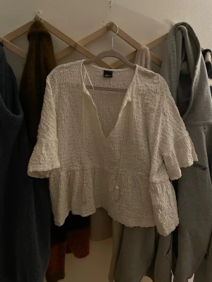 Vit strukturerad blus med volanger - Säljer en vit blus i krinklat tyg med v-ringning och knytband framtill. Blusen har volangdetaljer nedtill och vid ärmsluten, samt korta ärmar. Perfekt inför sommaren❤️