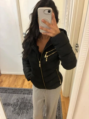 Svart pufferjacka med gulddragkedjor - Snygg svart pufferjacka med flera guldfärgade dragkedjor och fickor. Jackan har en figurnära passform och hög krage som skyddar mot kyla. Perfekt för kalla vinterdagar och ger en trendig look. Materialet är quiltat och ger extra värme.