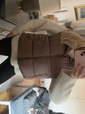 Brun/lila dunväst med hög krage - Snygg brun dunväst med hög krage och dragkedja framtill. Västen har två stora fickor på sidorna och quiltad design som ger en puffig look. Perfekt att slänga över en stickad tröja för extra värme. Materialet är mjukt och ger en avslappnad vibe.