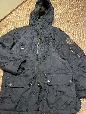 Mörkblå parkas med patch och huva - Säljer en mörkblå parkas med stor huva, flera fickor med knappar och dragkedjor samt en snygg patch med kompassmotiv på ärmen. Jackan har gulddetaljer på dragkedjorna och är tillverkad i slitstarkt syntetmaterial. Perfekt för kalla dagar och streetwear-stil.