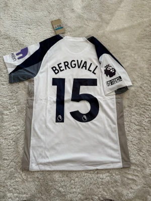 Tottenham matchtröja Bergvall 15 - Snygg Tottenham Hotspur matchtröja med Bergvall och nummer 15 på ryggen. Tröjan är vit med mörkblå och grå detaljer, Premier League-märke på ärmen och AIA som sponsor framtill. Tillverkad i lätt och ventilerande material, perfekt för fotboll eller träning.