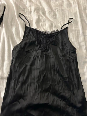 Svart linnetopp med spetsdetalj - Säljer en svart linnetopp med tunna axelband och v-ringning dekorerad med spets. Första bilden är framifrån och andra bilden bakifrån
