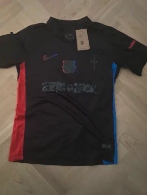Svart FC Barcelona x Nike t-shirt - Snygg svart t-shirt från Nike i samarbete med Travis scott.. Tröjan har Fc Barcelonas emblem i rött och blått, Nike-logga på bröstet och grafiskt tryck på magen. Röd och blå detalj på sidorna. Perfekt för dig som älskar fotboll och streetwear. 