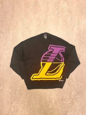 Svart knittedshirt NBA Boss  - Cool svart tröja med stort Los Angeles Lakers-logga i lila och gult framtill. Rund halsringning och långa ärmar. Perfekt för dig som älskar basket och vill sticka ut med en sportig stil.