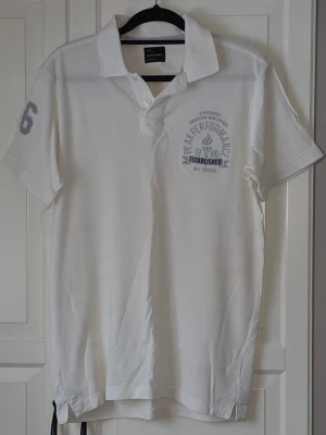 Vit Peak Performance pikétröja - Klassisk vit pikétröja från Peak Performance med tryck på bröstet och siffran 6 på ärmen. Den är i fint skick. Strl XL