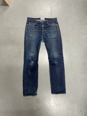 Mörkblå raka jeans från acne - Säljer ett par mörkblå jeans från Acne med raka ben och klassisk femficksmodell. Jeansen har snygga slitningar och kontrastsömmar, samt knappgylf. Perfekta för en avslappnad och stilren look.